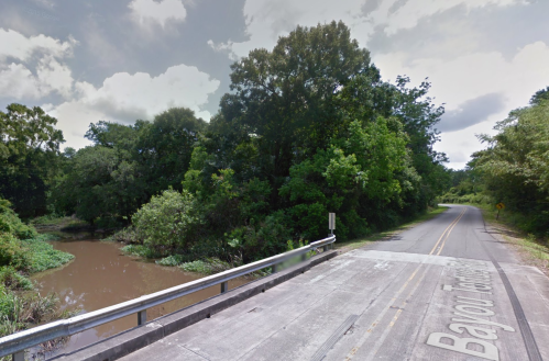 A quiet road alongside a river, surrounded by lush greenery and trees under a cloudy sky.