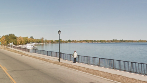 A person stands by a calm lake, with trees and boats in the background, along a paved walkway and street.