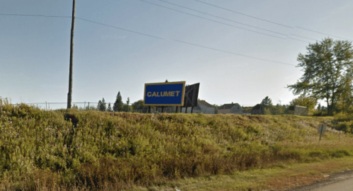 A roadside billboard for Calumet, surrounded by grass and trees under a clear sky.