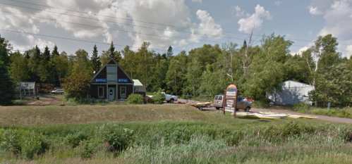 A small, triangular-roofed building with a sign, surrounded by trees and vehicles on a sunny day.