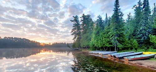 Serene lakeside scene at sunrise, with boats lined up on the shore and trees reflecting in the calm water.
