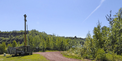 A drilling rig on a dirt path surrounded by green trees under a clear blue sky.