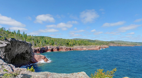 A scenic view of rocky cliffs and a blue ocean under a clear sky, surrounded by lush green trees.