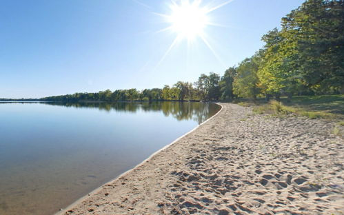 A serene lakeside view with a sandy shore, trees lining the water, and a bright sun shining in a clear blue sky.