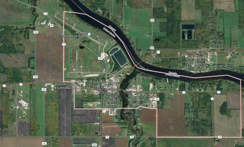 Aerial view of a small town near a river, surrounded by fields and roads, with a nearby airport visible.