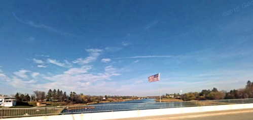 A wide view of a river under a blue sky, with a U.S. flag waving on a bridge in the foreground.