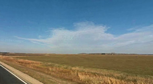 A wide-open landscape with grassy fields under a blue sky, featuring a distant horizon and a road on the left.