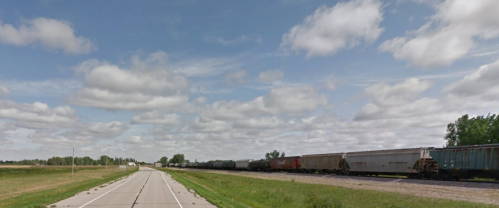 A wide view of a rural road alongside a train track with freight cars under a partly cloudy sky.