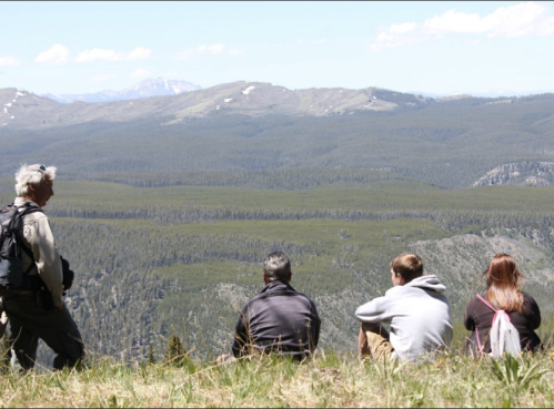 A group of four people sits on a grassy hill, overlooking a vast forested valley and distant mountains.