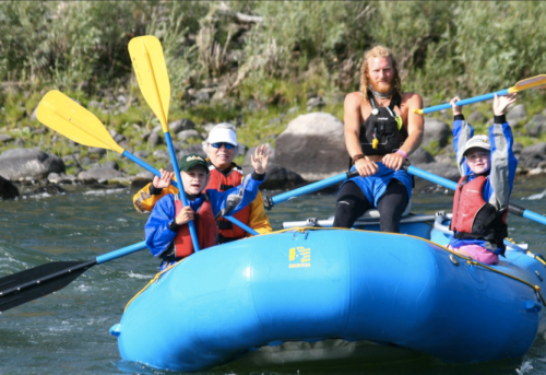 A group of four people in a blue raft on a river, smiling and waving, surrounded by greenery and rocky banks.