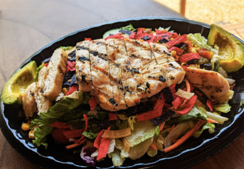 A colorful salad topped with grilled chicken, tortilla strips, and avocado, served in a black bowl.