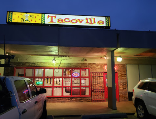 A brightly lit restaurant sign reading "Tacoville" at dusk, with an open door and nearby ATM.