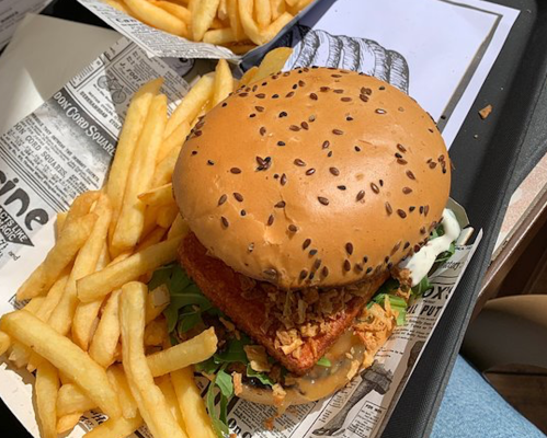 A large burger with a sesame seed bun, crispy chicken patty, lettuce, and fries on the side, served on newspaper print.