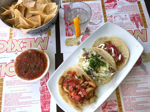 A plate of three tacos with toppings, a bowl of salsa, tortilla chips, and a drink on a colorful menu backdrop.
