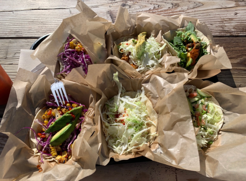 A tray of six tacos wrapped in paper, featuring colorful fillings like cabbage, corn, and lettuce on a wooden table.