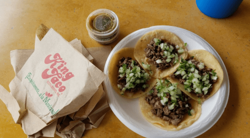 A plate of four tacos topped with meat and chopped onions, alongside a container of salsa and a takeout bag.