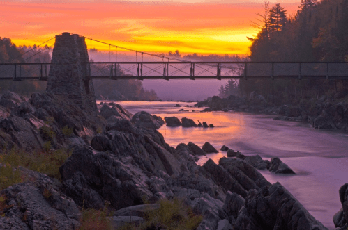 A scenic view of a bridge at sunset, with a river and rocky shoreline surrounded by trees and mist.