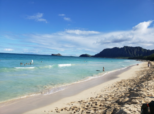 A scenic beach with clear blue water, gentle waves, and people enjoying the sun, framed by mountains in the background.