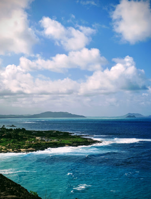 A scenic view of a coastline with turquoise waters, rocky shores, and fluffy clouds in a blue sky.