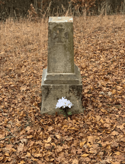 A weathered gravestone surrounded by fallen leaves, with white flowers placed at its base.
