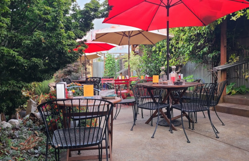 Outdoor dining area with wooden tables, black chairs, and red umbrellas surrounded by greenery.