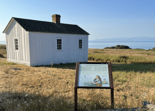 A historic white building with a sign about the Laundress Quarters, set against a scenic landscape and water view.