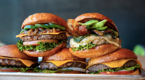 A close-up of four gourmet burgers stacked on a tray, featuring various toppings like cheese, bacon, and vegetables.