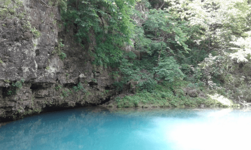 A serene blue lake surrounded by lush green trees and rocky cliffs, reflecting a peaceful natural landscape.