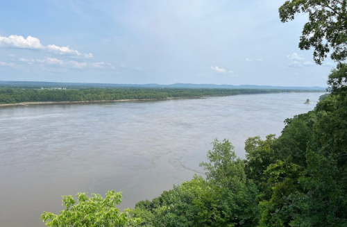 A wide river flows through a lush green landscape under a blue sky with scattered clouds.
