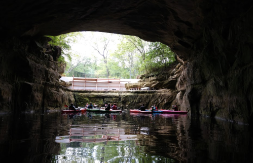 A group of kayakers paddles through a cave, surrounded by rocky walls and greenery, with light streaming in from above.
