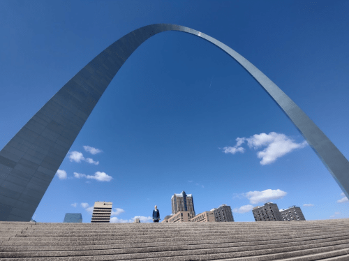 A person stands at the base of a large arch structure against a blue sky with clouds and city buildings in the background.