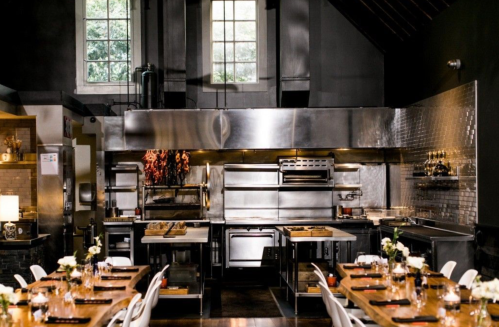 A modern kitchen with stainless steel appliances, wooden tables set for dining, and large windows letting in natural light.
