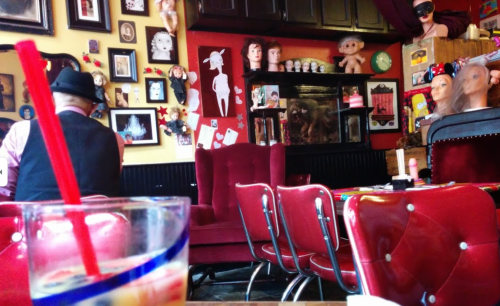 A quirky café interior with vintage decor, including dolls and masks, and a drink in the foreground.