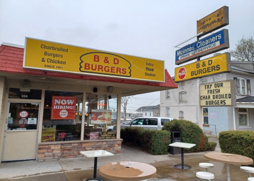 A burger restaurant with a bright sign, featuring "Now Hiring" and "Charbroiled Burgers & Chicken" in a rainy setting.