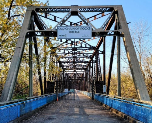 Old Chain of Rocks Bridge with a metal structure and a sign, surrounded by trees and a clear blue sky.