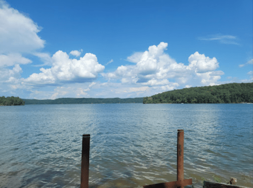A serene lake view with blue skies and fluffy clouds, surrounded by green hills and wooden posts in the foreground.