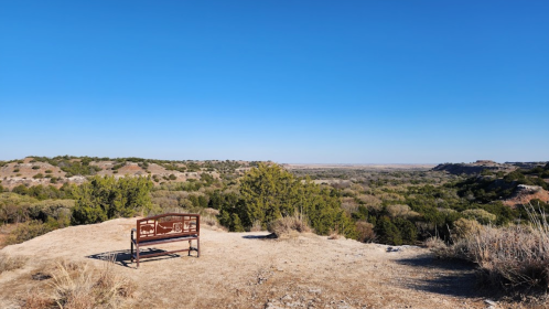A metal bench overlooks a vast, dry landscape with rolling hills and clear blue skies.