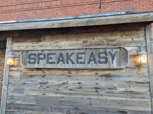 Wooden sign reading "SPEAKEASY" on a rustic wall, flanked by two lanterns.