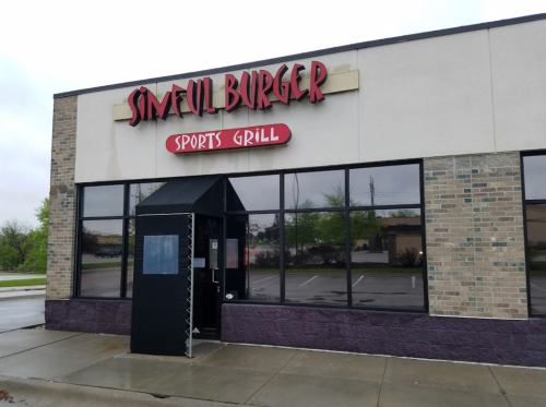 Exterior of Sinful Burger Sports Grill, featuring a sign and entrance, with a cloudy sky and wet pavement.