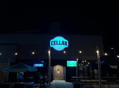 Exterior of "The Cellar" sports bar at night, featuring a lit sign and outdoor seating with string lights.