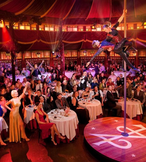 A lively circus performance with acrobats on a pole, surrounded by an engaged audience at elegantly set tables.