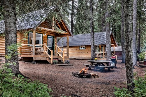 Two rustic log cabins nestled in a forest, with a picnic table and a campfire area surrounded by trees.