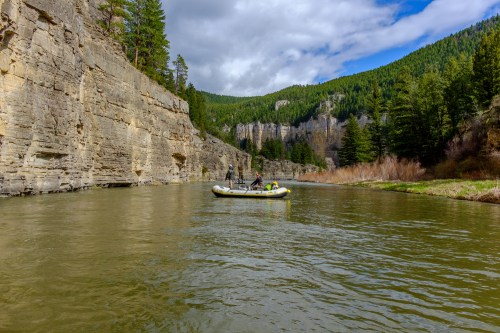 A small boat floats on a river surrounded by steep cliffs and lush green trees under a partly cloudy sky.