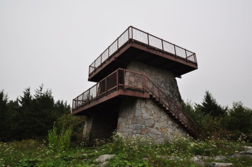 A stone observation tower with a wooden deck, surrounded by trees and wildflowers under a cloudy sky.
