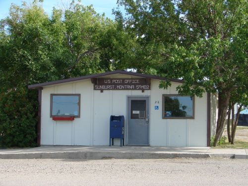 A small, single-story post office building in Sunburst, Montana, surrounded by trees and a gravel path.