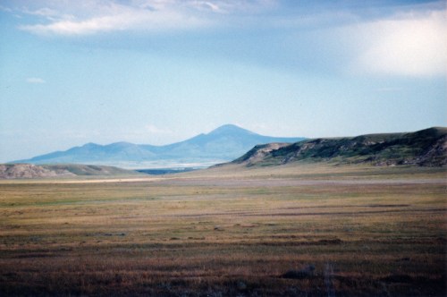 A vast, open landscape with rolling hills and a distant mountain under a blue sky with scattered clouds.