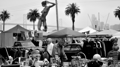 A bustling outdoor market with various items for sale, palm trees, and a large statue in the background.
