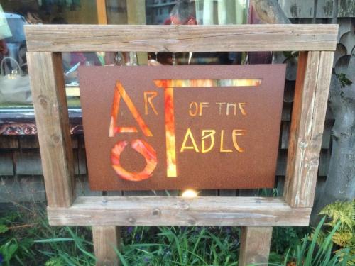 Sign for "Art of the Table" featuring stylized lettering on a rustic wooden frame, surrounded by greenery.