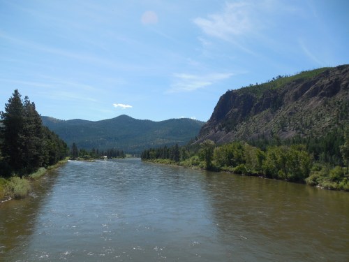 A serene river flows through a lush green landscape, surrounded by mountains under a clear blue sky.