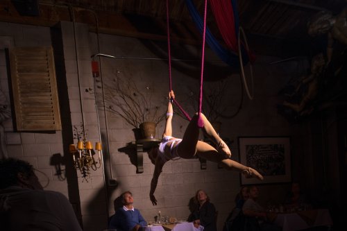 Aerial performer gracefully swings on pink silks above a dimly lit restaurant, with captivated diners below.